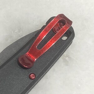 Pu&ograve; includere: Primo piano di un coltello pieghevole nero con una clip da tasca rosso brillante. La clip &egrave; fissata con due piccole viti e ha un design curvo. L'impugnatura del coltello &egrave; strutturata e una singola vite rossa &egrave; visibile vicino alla base della clip.