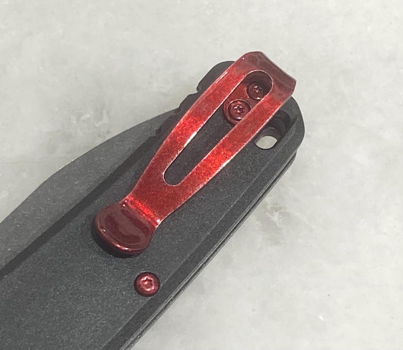 Pu&ograve; includere: Primo piano di un coltello pieghevole nero con una clip da tasca rosso brillante. La clip &egrave; fissata con due piccole viti e ha un design curvo. L'impugnatura del coltello &egrave; strutturata e una singola vite rossa &egrave; visibile vicino alla base della clip.