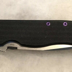 May include: A black folding knife with a textured handle and a silver blade. The knife has five purple screws on the handle. The handle is marked with the number "3000".