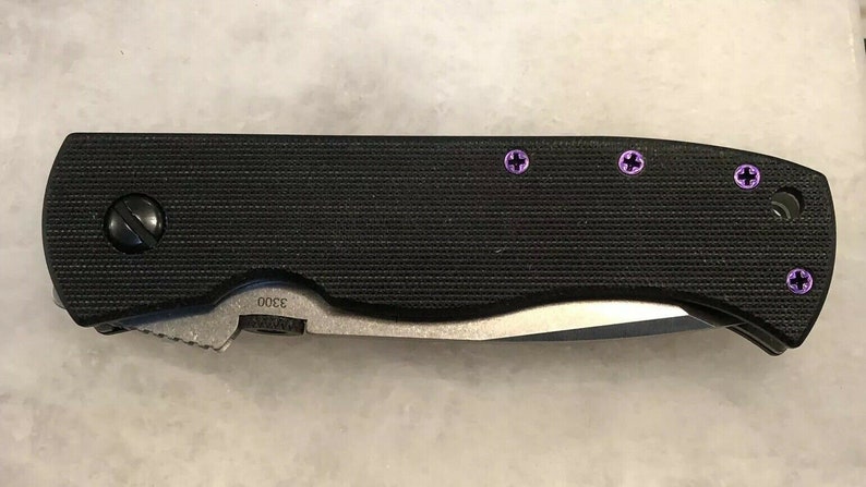 May include: A black folding knife with a textured handle and a silver blade. The knife has five purple screws on the handle. The handle is marked with the number "3000".