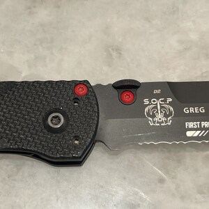 May include: A black folding knife with a serrated blade and a black handle. The knife has a red button on the side and the text "S.O.C.P. D2 GREG THOMSON FIRST PRODUCTION 788" is engraved on the blade.