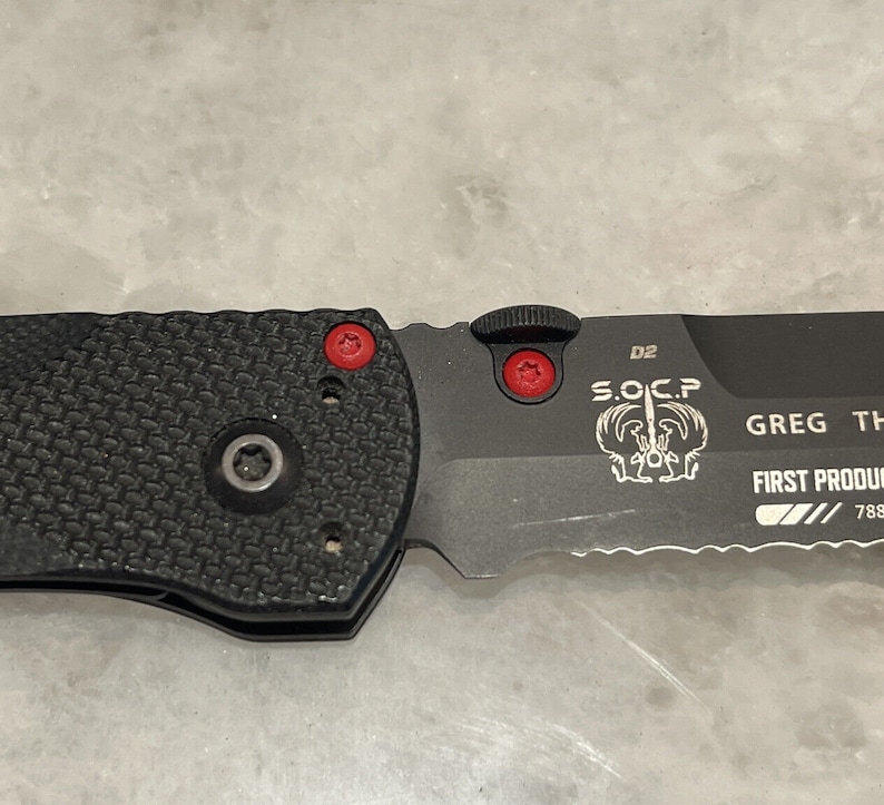 May include: A black folding knife with a serrated blade and a black handle. The knife has a red button on the side and the text "S.O.C.P. D2 GREG THOMSON FIRST PRODUCTION 788" is engraved on the blade.
