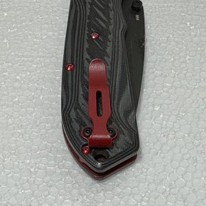 Puede incluir: Una navaja plegable con un mango texturizado negro y gris y una hoja negra. El cuchillo presenta detalles en rojo en el clip de bolsillo y los tornillos. La hoja está marcada con "M4".