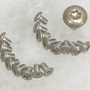 May include: A collection of small, silver-colored screws with a star-shaped indentation on the head. One larger, brass-colored screw is also present. The screws are arranged in curved lines, suggesting a hardware or repair theme.