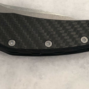 以下が含まれることがあります： 黒いカーボンファイバー製のナイフのハンドルをクローズアップ。銀色のネジが4つ付いています。ハンドルにはテクスチャ加工が施されています。