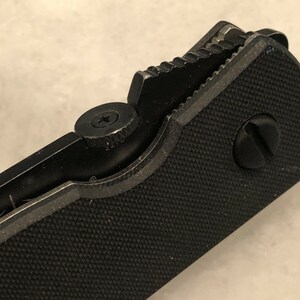 May include: Close-up of a black folding knife with a textured handle and a partially open blade. The knife has a screw on the handle and a serrated edge on the blade.