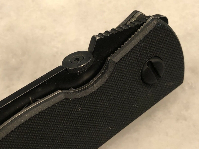 May include: Close-up of a black folding knife with a textured handle and a partially open blade. The knife has a screw on the handle and a serrated edge on the blade.