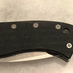 May include: Close-up of a black pocket knife handle with four silver screws. The handle has a textured surface.