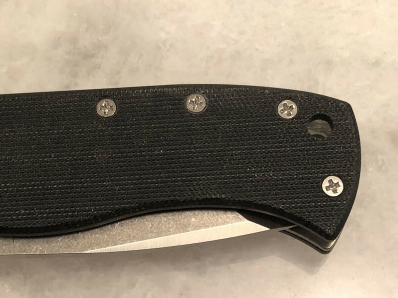 May include: Close-up of a black pocket knife handle with four silver screws. The handle has a textured surface.