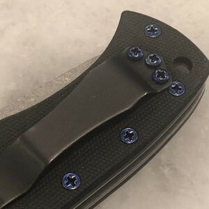 May include: Close-up of a black pocket knife with a blue titanium clip and blue screws. The knife has a black handle and a silver blade.