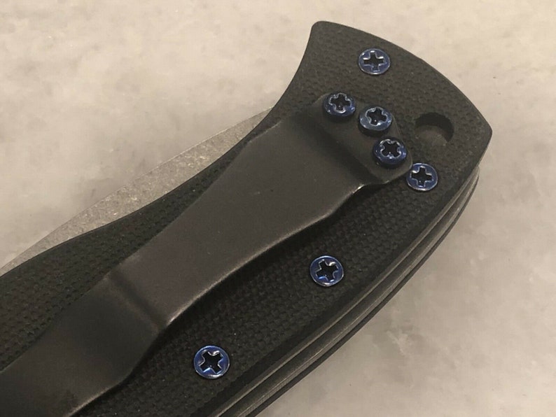 May include: Close-up of a black pocket knife with a blue titanium clip and blue screws. The knife has a black handle and a silver blade.