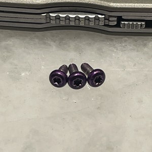 May include: Three purple screws with star-shaped heads are arranged on a light-colored surface. The screws have a metallic sheen and are positioned near a multi-tool. The screws are small, likely for precision work.