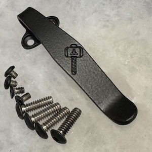 May include: A black metal pocket clip with a hammer and anvil logo. The clip is surrounded by eight silver screws.