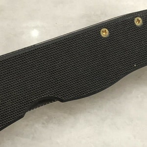 May include: Close-up of a black folding knife with a textured handle and gold screws. The knife is closed and the blade is not visible.
