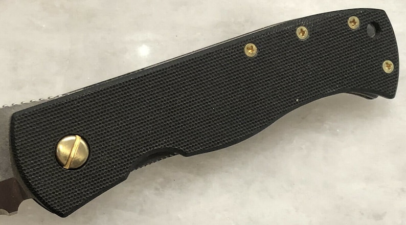 May include: Close-up of a black folding knife with a textured handle and gold screws. The knife is closed and the blade is not visible.