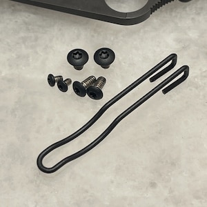Könnte beinhalten: Ein schwarzer Metall-Taschenclip mit zwei Schlaufen und fünf schwarzen Schrauben.