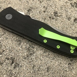 Pode incluir: Uma faca dobr&aacute;vel preta com um clipe de bolso verde. A faca tem uma al&ccedil;a preta texturizada e uma l&acirc;mina prateada.