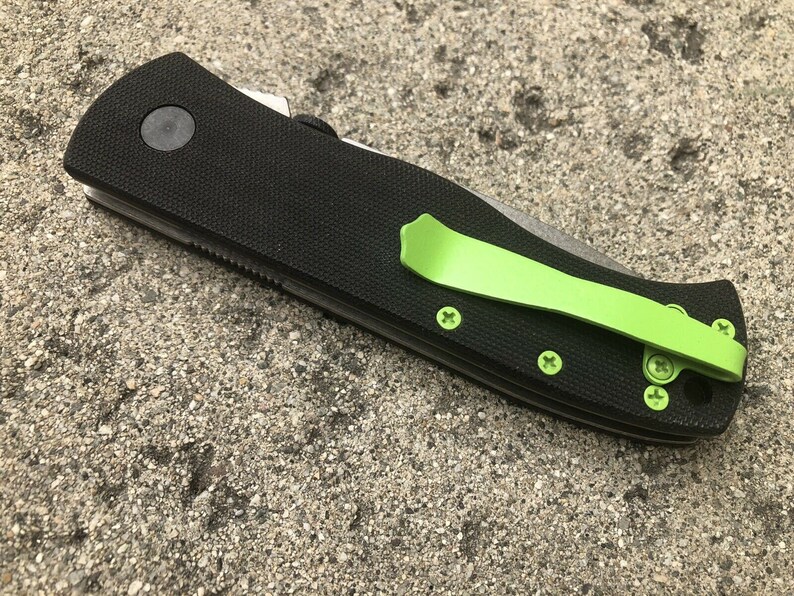 Pode incluir: Uma faca dobr&aacute;vel preta com um clipe de bolso verde. A faca tem uma al&ccedil;a preta texturizada e uma l&acirc;mina prateada.