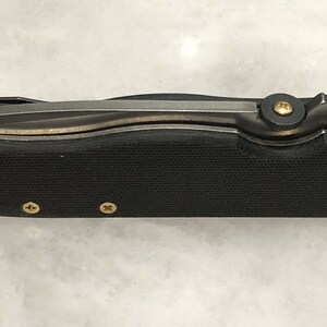 May include: A black folding knife with a textured handle and gold accents. The knife is closed and has a partial view of the blade.
