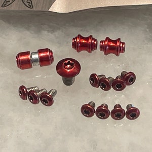 Op de afbeelding: Een verzameling rode metalen hardware, waaronder schroeven, bouten en tonvormige stukken. De hardware is gerangschikt op een wit oppervlak.
