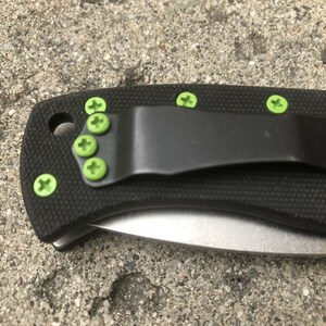 May include: A black folding knife with a green pocket clip and green screws. The knife has a black handle and a silver blade.