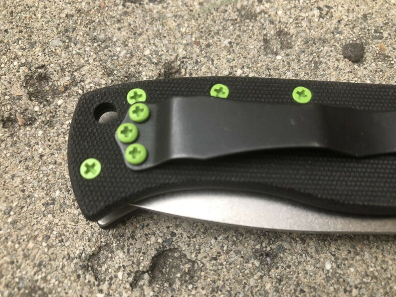 May include: A black folding knife with a green pocket clip and green screws. The knife has a black handle and a silver blade.
