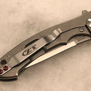 May include: A silver pocket knife with a black blade and a red pocket clip. The knife has a ZT logo on the clip and the text "0450 CLA  S30V  USA" on the handle.