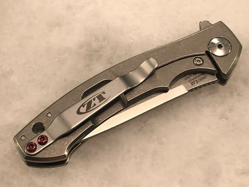 May include: A silver pocket knife with a black blade and a red pocket clip. The knife has a ZT logo on the clip and the text "0450 CLA  S30V  USA" on the handle.