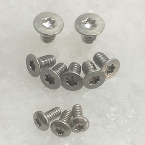 May include: A collection of nine silver-colored metal screws with flat heads and star-shaped Torx drive recesses. The screws are arranged in three rows, featuring threaded shanks and a metallic finish, designed for secure fastening applications.