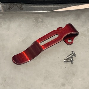 May include: A red metal pocket clip with two silver screws. The clip is curved and has a smooth finish.