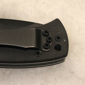 May include: Close-up of a black pocket knife with a clip. The clip is attached to the knife with five screws.