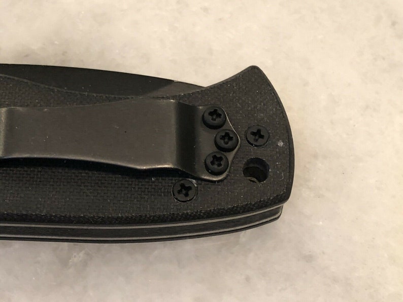 May include: Close-up of a black pocket knife with a clip. The clip is attached to the knife with five screws.