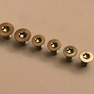 Puede incluir: Seis tornillos plateados con un hueco en forma de estrella en la cabeza. Los tornillos están dispuestos en diagonal, con las cabezas visibles. Las cabezas son circulares. Para mecánica o construcción.