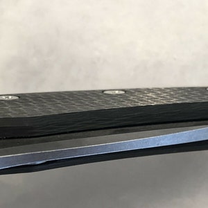 以下が含まれることがあります： 折りたたみナイフの黒いカーボンファイバーハンドルと銀色のネジのクローズアップ。