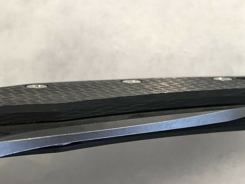 以下が含まれることがあります： 折りたたみナイフの黒いカーボンファイバーハンドルと銀色のネジのクローズアップ。