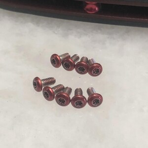 May include: A collection of eight small metal screws with rounded, metallic red heads and silver-colored threaded shanks. Each screw head features a star-shaped torx drive pattern in the center, arranged in two rows on a light, textured surface.