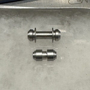 May include: Two silver metal axles, one longer than the other, with a cylindrical shape and a grooved design on the ends.