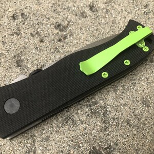 Pode incluir: Uma faca dobr&aacute;vel preta com um clip de bolso verde. A faca tem uma al&ccedil;a texturizada e uma l&acirc;mina preta.