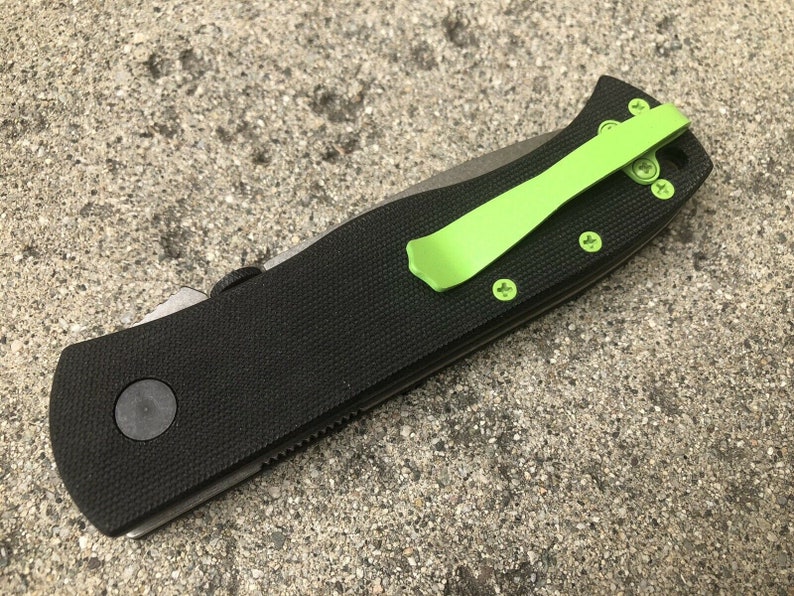 Pode incluir: Uma faca dobr&aacute;vel preta com um clip de bolso verde. A faca tem uma al&ccedil;a texturizada e uma l&acirc;mina preta.