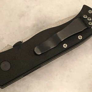 May include: Black folding knife with a black pocket clip and a textured black handle. The knife has a silver blade and a silver pocket clip.