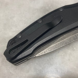 May include: A black folding knife with a partially open blade. The knife has a black handle with a textured finish and a pocket clip.