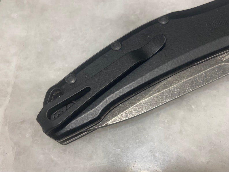 May include: A black folding knife with a partially open blade. The knife has a black handle with a textured finish and a pocket clip.
