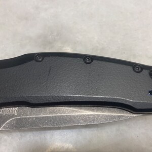 May include: A black folding knife with a silver blade and a black handle. The handle has four screws and a small hole near the blade.