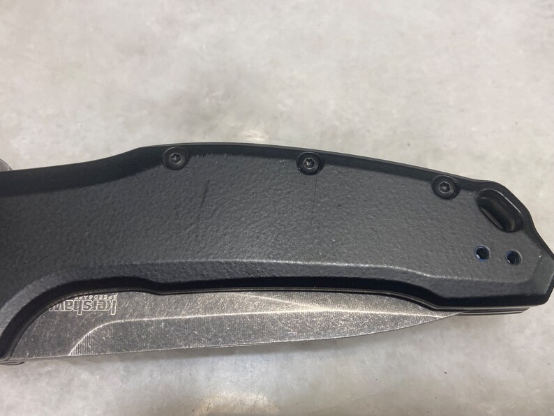 May include: A black folding knife with a silver blade and a black handle. The handle has four screws and a small hole near the blade.