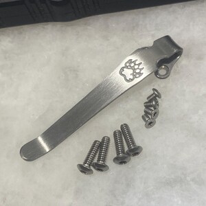 May include: A silver pocket clip with a bear paw print design. The clip is shown with six silver screws.