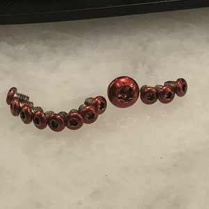 May include: A close-up of a collection of small, red, star-shaped screws. The screws are arranged in a semi-circle with a larger, red, star-shaped screw in the centre.