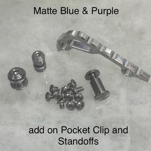 Puede incluir: Un conjunto de clip de bolsillo y separadores de metal que incluye un clip largo texturizado, dos separadores redondeados, un separador cilíndrico y nueve tornillos pequeños. El texto Matte Blue & Purple y add on Pocket Clip and Standoffs es visible.