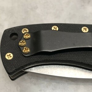 May include: A black folding knife with a textured handle and gold screws. The knife has a pocket clip.