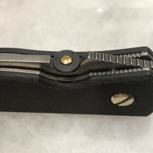 May include: Close-up of a black pocket knife with a textured handle and a silver blade. The knife has a gold screw on the handle and a gold screw on the blade.