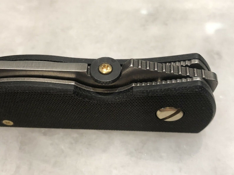 May include: Close-up of a black pocket knife with a textured handle and a silver blade. The knife has a gold screw on the handle and a gold screw on the blade.
